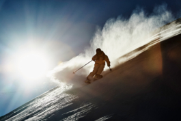 Skier coming towards the camera off piste with the sun shinning behind. With a bloom of snow, rooster blowing uphill. Ross Woodhall - Action & Lifestyle Photography