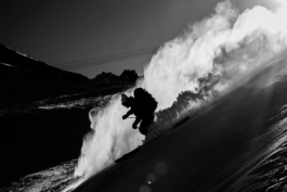 Black and white image of skier with backpack and wooly hat on skiing down hill with blooms of snow hightlighted by the sun. Ross Woodhall - Action & Lifestyle Photography