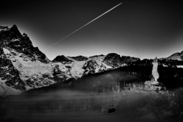 Black and white image of a mountain with a telecabine. in the sky is a bright white aeroplane contrails. Vapour trail. Le Meije. In the Massif des écrins range Ross Woodhall - Action & Lifestyle Photography