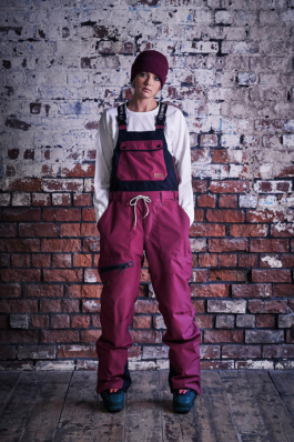 Woman wearing burgundy coloured ski dungarees with matching hat with white long sleeved shirt in front of a whitewashed brick wall. Ross Woodhall - Action & Lifestyle Photography