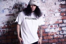 Man with long fuzzy hair wearing Planks clothing t shirt and cap leaning against a whitewashed brick wall with tattooed arms. Ross Woodhall - Action & Lifestyle Photography