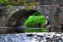 Man fly fishing by a brick bridge, wearing waders and in the water, caught mid cast, with a fishing net hooked to his backpack. Ross Woodhall - Action & Lifestyle Photography