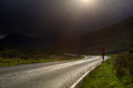 Male runner jogging along a windy road surrounded by rough grass and ferns with moody dark clouds with hills in the background. Ross Woodhall - Action & Lifestyle Photography