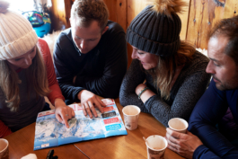 four skiers having a hot chocolate looking at a piste map, the two women are wearing their wooly hats. Ross Woodhall - Action & Lifestyle Photography