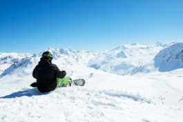 Single snowboarder sat on the slope looking out at the mountains and slope in the background with people on. Ross Woodhall - Action & Lifestyle Photography