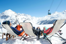 three skiers having a relax in sun lounger chairs, with a telecabine in the background, surrounded by mountains. Ross Woodhall - Action & Lifestyle Photography