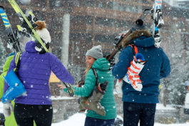 four skiers walking with their skis and boots over their shoulders, wearing jackets and wooly hats whilst it is snowing. Ross Woodhall - Action & Lifestyle Photography