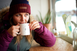 woman sat having a hot drink from a mug, wearing a puffer coat and wooly hat with skis propped up against the window outside. Ross Woodhall - Action & Lifestyle Photography