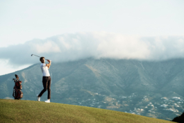 Man playing golf, with golf bag beside him, having hit the ball. Mountain in the background, with the tip of the mountain hidden behind clouds. Houses/building seen at lower level of the mountain. Ross Woodhall - Action & Lifestyle Photography