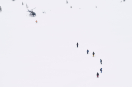 A snowy landscaoe taken from an angle above. A group of 7 skiers, snowboarders making their way back to a helicopter. Ross Woodhall - Action & Lifestyle Photography