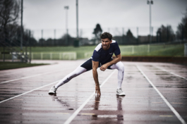 Man stretching on a wet running track, wearing white leggings underneath blue shorts with a blue and white top. Both clothes are Castore branding. Ross Woodhall - Action & Lifestyle Photography