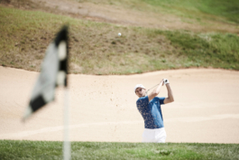 Man playing golf, wearing Castore clothing. Hitting the ball towards the green from a sand bunker. Flag in foreground. Ross Woodhall - Action & Lifestyle Photography
