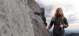 Woman with red hair looking out into the distance against a rocky cliff side with hills also in the background. Wearing a black lightweight quilted jacket, carrying a water bottle from Land Rover, Musto brand. Ross Woodhall - Action & Lifestyle Photography