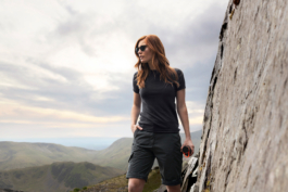 Woman with red hair looking out into the distance against a rocky cliff side with hills also in the background. Wearing a short dark grey t shirt & shorts, carrying a water bottle from Land Rover, Musto brand. Wearing sunglasses. Ross Woodhall - Action & Lifestyle Photography