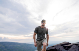 Man walking on a hill top with hills in the distance. Wearing a short sleeved t shirt & shorts with a cap & sunglasses. You can't see the mans feet or boots. Clouds in the sky with the sun still bright in the sky directly behind him. Ross Woodhall - Action & Lifestyle Photography