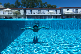 two thirds of image is a woman in crystal clear water, shot underwater with arms out, last third, top of image is the poolside umbrellas and hotel apartments. Ross Woodhall - Action & Lifestyle Photography