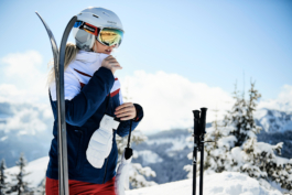 female skier zipping her jacket up, with skis and poles next to her, with mountains in the background. Ross Woodhall - Action & Lifestyle Photography