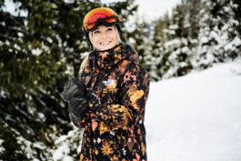 female skier with winter jacket, gloves, helmet with googles on, pine trees in the background. Ross Woodhall - Action & Lifestyle Photography