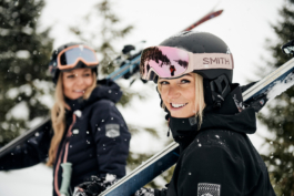 two female skiers walking with their skis over their shoulders, nearest female looking towards the camera. Ross Woodhall - Action & Lifestyle Photography