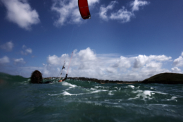 Kite surfer in the sea with red sail in the air just about to set off with land in the background. Ross Woodhall - Action & Lifestyle Photography