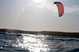 Kite surfer, seen with red sail. Taken from the water with land in the background. Ross Woodhall - Action & Lifestyle Photography