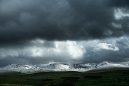 Snowy peaks with green dark ground in foreground with moody clouds and sun light on to the snow. Ross Woodhall - Action & Lifestyle Photography