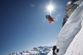 Snowboarder jumping off a cliff, over a cornice, grabbing his board with one hand. The sun directly above him. Clear blue skies. Ross Woodhall - Action & Lifestyle Photography