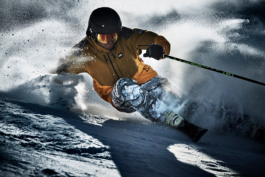 Solo skier powering off piste through the snow. Wearing a black helmet with reflective googles, mustard coloured jacket & patterned white and grey ski pants. Snow flying from his ski's. Ross Woodhall - Action & Lifestyle Photography