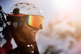 Bearded man wearing a white ski helmet, mirrored googles and a small view of his ski's over his shoulders, smiling looking out. Ross Woodhall - Action & Lifestyle Photography