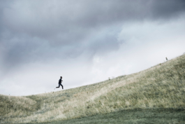 Man running slightly up hill over some rough grassed area with cloudy sky. Ross Woodhall - Action & Lifestyle Photography