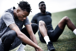 Two black/ethnic/coloured men resting after running. one sat on the ground with legs bent out in front of him, the other is squating with beats of sweat running off his face. Ross Woodhall - Action & Lifestyle Photography