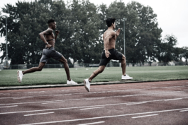 Two men on running track, both sprinting and mid air. Both topless, one wearing heart monitor. Large trees in background. Ross Woodhall - Action & Lifestyle Photography