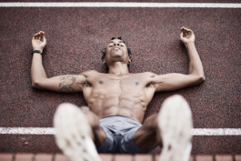 Man on running track, topless with legs up against a wall, looking towards the camera, between his feet. Ross Woodhall - Action & Lifestyle Photography