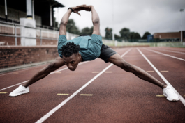 Black/ethnic/coloured man on running track, stretching with legs spread wide, head down and arms pushed back and raised. Wearing Castore branded T shirt and shorts. Ross Woodhall - Action & Lifestyle Photography