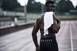 Black man topless on running track, with T shirt over his shoulder, looking back towards the camera, he is wearing a running monitor and has Castore branding on his shorts. Ross Woodhall - Action & Lifestyle Photography