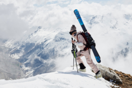 woman skier, with skis strapped to her backpack, with gloves hanging from her hands. Mountains and clouds in the background. Ross Woodhall - Action & Lifestyle Photography