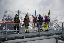 five skiers with their skis strapped to their backpacks, on a high metal bridge, looking at the view and mountains in the background. Ross Woodhall - Action & Lifestyle Photography
