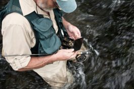 Man fly fishing looking at his box of flies, wearing a fishermans gilet jacket and waders standing in water up to his knees. Ross Woodhall - Action & Lifestyle Photography