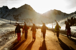 6 back country skiers with backpacks on, carrying ski's on their shoulders, walking away from the camera abreast to each other. Walking down a winding tarmaced road with snow covered ground each side with guide poles for the road side. Golden glow from the sun soon to set behind the mountain range. Ross Woodhall - Action & Lifestyle Photography