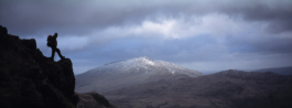 single hiker silhouette with a backpack on, stood on a ridge whilst looking out towards a snow capped hill in the distance. Ross Woodhall - Action & Lifestyle Photography