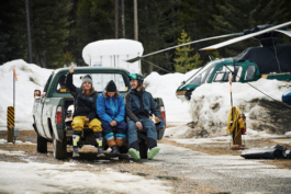Three backcountry skiers sat in ski clothing on the back of a truck with their legs dangling. Helicopter parked in the background. Ross Woodhall - Action & Lifestyle Photography