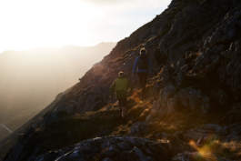 Two hikers, walking up a hill with rocks and rough grass with a shaft of sunlight. Ross Woodhall - Action & Lifestyle Photography