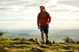 Man hiking/walking across rough ground, wearing shorts and light jacket and a backpack. views on more green fields below him and in the distance. Ross Woodhall - Action & Lifestyle Photography