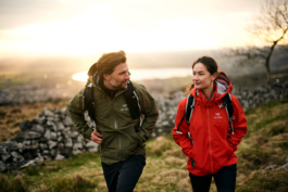 Man and woman out walking/hiking. Wearing Arc.Teryx jackets with backpacks. Walking by a stonewall with a lake in the distance behind them. Ross Woodhall - Action & Lifestyle Photography