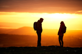 Two hikers, a man and a woman paused from walking/hiking. Chatting, facing each other with the sunsetting, with different shades of orange and shadows of hills seen in the distance. Wearing backpacks. Ross Woodhall - Action & Lifestyle Photography