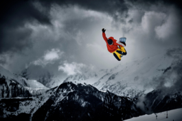 Snowboarder in the air, grabbing his board with other arm straight up into the air. He is wearing bright red jacket with white strip down the arms with yellow & black trousers. He is bright in the image, a contrast against the moody black & white background of mountains & clouds. Ross Woodhall - Action & Lifestyle Photography