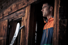 male skier, standing in wooden chalet doorway with pair of skis propped up against the wall. Ross Woodhall - Action & Lifestyle Photography