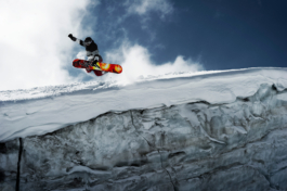 Solo snowboarder jumping from a glacier wall off piste. Grabbing his snowboard with one hand & showing his bright snowboard against the grey glacier ice wall. Ross Woodhall - Action & Lifestyle Photography