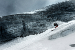 Solo skier skiing off piste along & over a glacier where behind him is a large slab of glacier ice exposed showing lines in the ice. Image is staturated. Ross Woodhall - Action & Lifestyle Photography