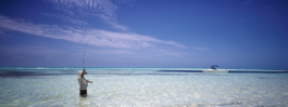 Man up to his thighs in crystal clear sea water in the Caribbean fly fishing with small motor boat anchored further out. Ross Woodhall - Action & Lifestyle Photography